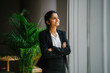 © Danon - Portrait of a young Asian Indian professional woman in a meeting room standing by the glass window, smiling with her arms crossed. She looks optimistic, happy and confident