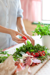 Wall Mural - Woman is cooking in home kitchen. Female graceful hands hold cherry tomatoes. Ingredients for preparing italian or french food are on table on wooden boards. Lifestyle moment. Close up.