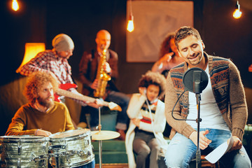 Poster - Man singing and sitting on chair while his band playing instruments in background. Home studio interior.