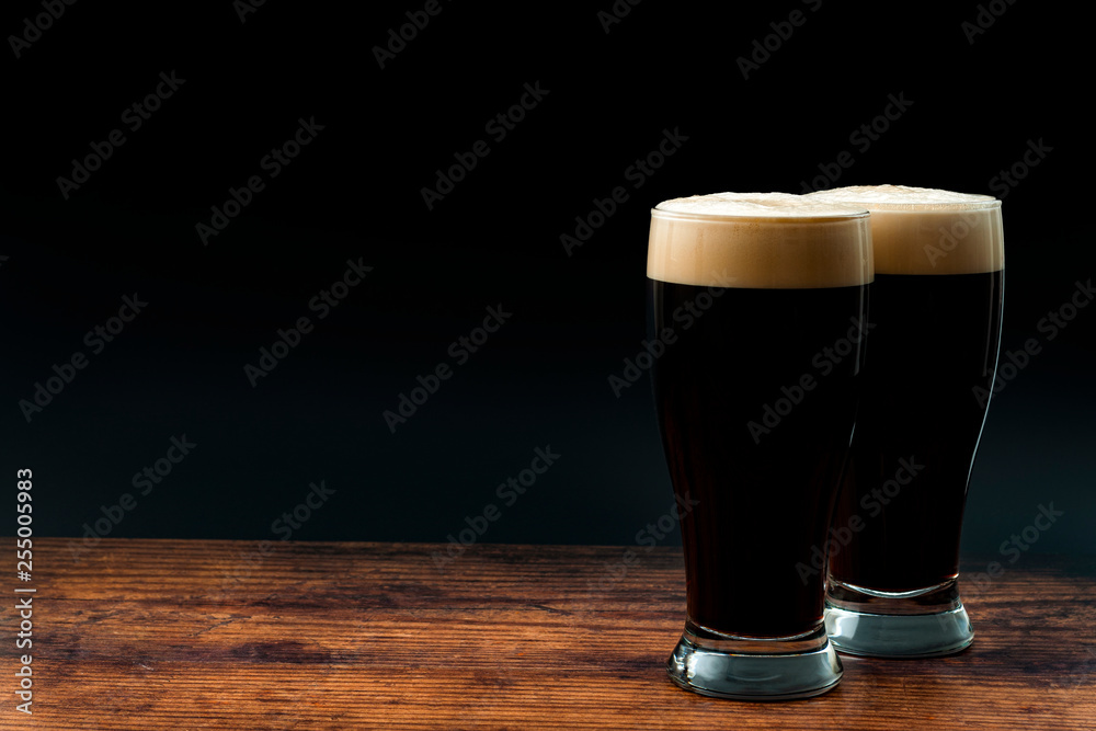Alcohol abuse, bitter refreshing alcoholic beverage and dry stout concept theme with frothy glass pints of dark beer on wood table isolated on black background with copyspace in bar or pub setting - obrazy, fototapety, plakaty 