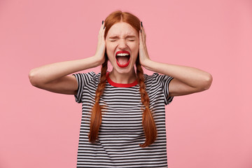 Wall Mural - Angry young red-haired girl in stripped t-shirt, keeps closed eyes, closes her ears with palms, showing gesture of ignore, demonstrates loud scream or shout, over pink wall