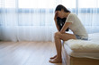 © PR Image Factory - sad asian woman suffering from depression sit on bed hands on forehead. depressed young girl in shorts in bedroom on wooden floor next to sunshine window with curtain. upset lady indoors thinking.