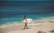 © South House Studio - slim young surfer woman carries surfboard under her arm, walking on pink sand