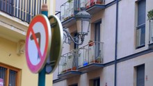 Facades Of Houses On A Narrow Street In The City Of Spain, On The Wall Hangs An Ancient Street Lamp And A Road Sign Prohibiting Traffic To The Left. Shot In Motion