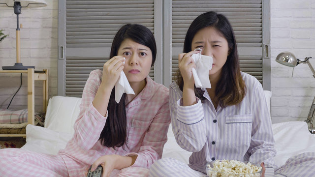 two chinese best friends watch sad movie at home in bed crying touching. girls hold popcorn and tv remote control looking screen romantic film on television. ladies in pajama wiping tear using tissue