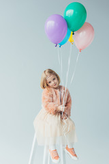 Wall Mural - adorable kid in faux fur coat and skirt sitting on highchair, looking at camera and holding balloons isolated on grey