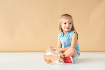 Wall Mural - cute kid sitting on floor with fishbowl and looking away on beige background