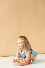 Wall Mural - cute kid sitting with crossed legs and holding fishbowl on beige background