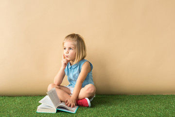 Wall Mural -  cute child sitting with crossed legs on grass rug and holding book on beige background