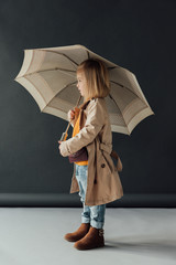 Wall Mural - side view of child in trench coat and jeans holding umbrella on black background