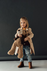 Wall Mural - smiling and cute child in trench coat and jeans holding teddy bear on black background