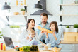 © Iryna - Happy young family with Mum, Dad and two different age children cooking breakfast, laughing biting the baguette in the kitchen preparing meal together.