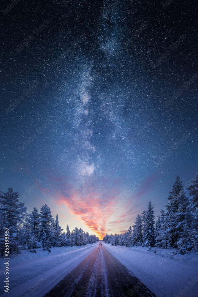 Road leading towards colorful sunrise between snow covered trees with epic milky way on the sky - obrazy, fototapety, plakaty 