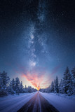 Fototapeta Natura - Road leading towards colorful sunrise between snow covered trees with epic milky way on the sky