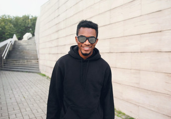 Wall Mural - Portrait happy smiling african man in black hoodie, sunglasses on city street over gray brick wall background