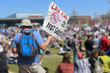 Back view of man in shorts and teeshirt with hat and backpack at gun control rally with a sign that says LOVE KIDS - Ban ARs not guns in front of blurred out crowd and stadium
