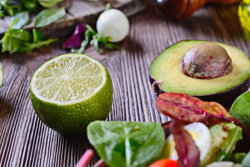 Half a ripe and healthy avocado with a bone next to half a lime and greens for a salad on a wooden table