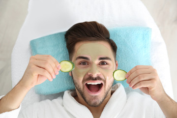 Wall Mural - Young man with clay mask on his face holding cucumber slices in spa salon, above view