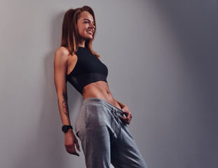 Fashion, sport, health. Slim fitness girl wearing sportswear leaning on a wall in a studio. Isolated on a gray background