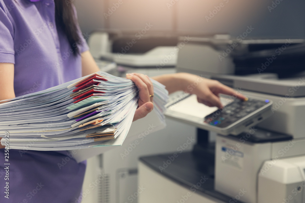 Bussinesswoman using copier machine to copy heap of paperwork in office. - obrazy, fototapety, plakaty 