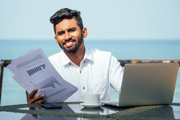 Wall Mural - handsome and successful indian man in a stylish well-dressed freelancer working with a laptop on the beach.freelance and remote work.businessman student in a summer cafe on the shore of india ocean