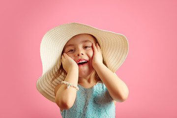 Wall Mural - Portrait of surprised girl with open moutn, wears beach hat and beautiful blue dress, expresses joy and happiness, stands over pink isolated background.