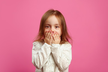 Little girl with a surprised look covered her mouth with hands and looks at you on pink isolated background.