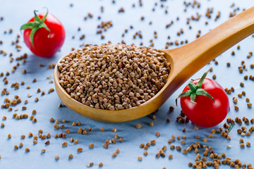 Dry buckwheat in wooden spoon and fresh ripe tomatoes cherry on blue background. Buckwheat cereal grains for dietary, slimming and healthy food