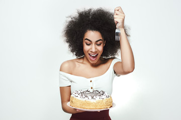 I will eat it right now! Excited cute afro american woman with curly hair is holding a fork in one hand and birthday cake in another, going to eat some piece of cake.