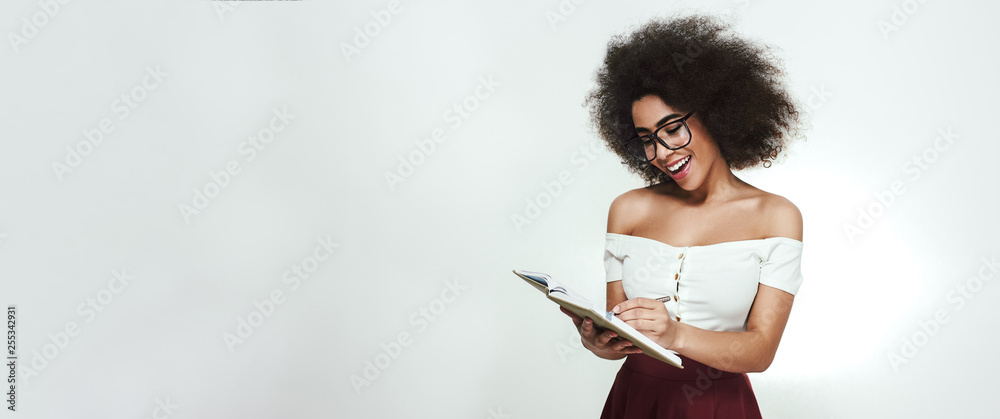 Planning her day. Cheerful and young african woman in eyewear writing something in her notebook while standing against grey background - obrazy, fototapety, plakaty 