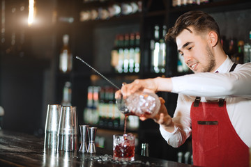 Smiling friendly looking barman wearing the bow tie, white shirt and red apron makes cocktail at bar counter at restaurant