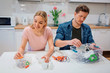 © estradaanton - Recycling, reuse, energy. Young couple sorting batteries, other electronic waste into containers with recycling symbol while sitting at kitchen