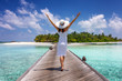 Glückliche Frau in weißem Sommerkleid läuft über einen Holzsteg auf den Malediven und genießt ihren tropischen Uraub