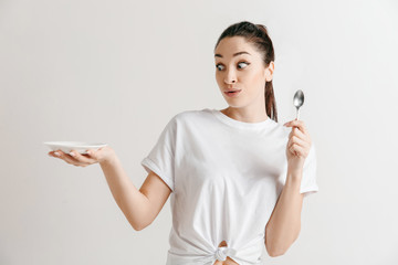 Young fun crazy brunette housewife with spoon isolated on white background. Housekeeper woman holding white empty plate. Copy space advertisement.