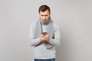 Puzzled young man in gray sweater, scarf using mobile phone typing sms message isolated on grey background. Healthy fashion lifestyle, people sincere emotions, cold season concept. Mock up copy space.