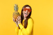 Crazy young woman in funny glasses showing tongue, holding in hands fresh ripe pineapple fruit isolated on yellow orange background. People vivid lifestyle relax vacation concept. Mock up copy space.