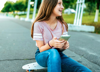 A little girl schoolgirl of 12-16 years old, on street she sits on a skateboard in summer in park. In hands of phone, online application, Internet social networks, smiling rejoices and laughs happy.