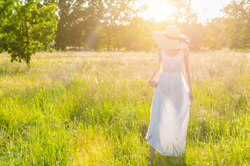 Wall Mural - Happy beautiful positive fun smiling girl woman wear straw hat long white dress shawl chiffon fabric walking step on big large wide summer hot spring sunny green sunray light field between fruit trees