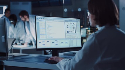 Fototapete - Over the Shoulder Shot: Female IT Scientist Uses Computer Showing System Monitoring and Controlling Program. In the Background Technology Development Laboratory with Scientists, Engineers Working 