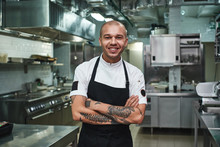 I Love My Work Cheerful Young Chef In Apron Keeping Tattooed Arms Crossed And Smiling While Standing In A Restaurant Kitchen