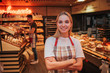 Happy young woman stand in grocery store. She pose nad look on camera. Happy worker smiling. Young man work behind. He put bread on shelf.