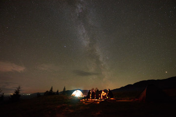 Summer night camping under stars. Group of five people, men and woman with guitar sitting by burning campfire at tourist tents under dark sky with bright sparkling stars and Milky Way constellation.