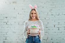 Happy Easter!Portrait Of Young Beautiful Woman With Bunny Ears Standing Near White Wall And Holding Basket Of Eggs.