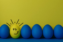Five Blue And One Yellow Funny Faced Painted Easter Eggs Stand In A Row On A Yellow With Blue Background. Happy Easter Holiday Card Or Banner. Copy Space.