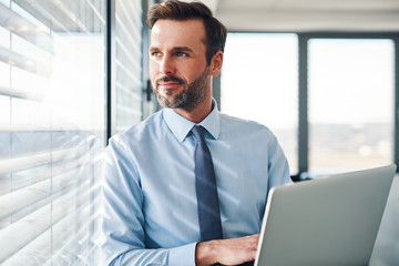 Sticker - Handsome businessman standing with laptop, looking throught the window in modern office