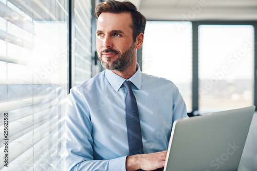 Fotomural Handsome businessman standing with laptop, looking throught the window in modern