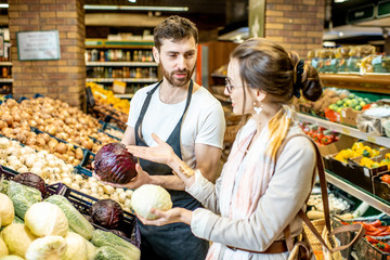 Shop worker helping young woman client to chooose vegetables in the supermarket