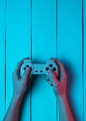 Wall Mural - Concept gamer. Nightlife. Leisure. Hand holds gamepad on blue wooden background. Neon red blue light. Retro style. Top view