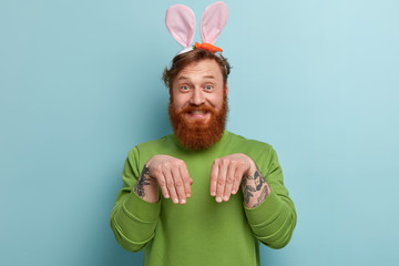 Wall Mural - Funny positve man with ginger beard, haircut, keeps hands as if rabbit, wears soft bunny ears, has friendly look, smiles cheerfully, ready to congratulate children with coming Easter. I am lovely hare