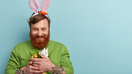 Wall Mural - Good looking optimistic redhaired guy shows Easter symbols small fluffy white rabbit, painted eggs in green grass, wears bunny ears on head, enjoys mystery of religious holiday. Mock up space aside